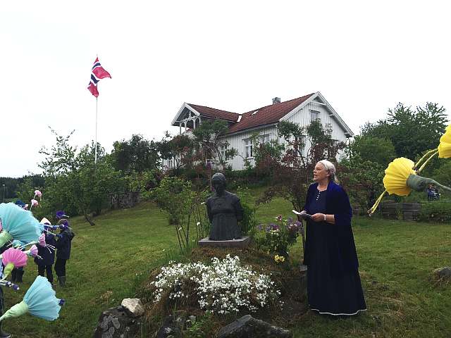 Sveitservillaen Fredheim i bakgrunnen med flagg på flaggstanga. Gunnar Janssons skulptur av den unge Ingeborg Refling Hagen i halvfigur i forgrunnen, Dagne Groven Myhren i blå kjole til høyre, talende.
